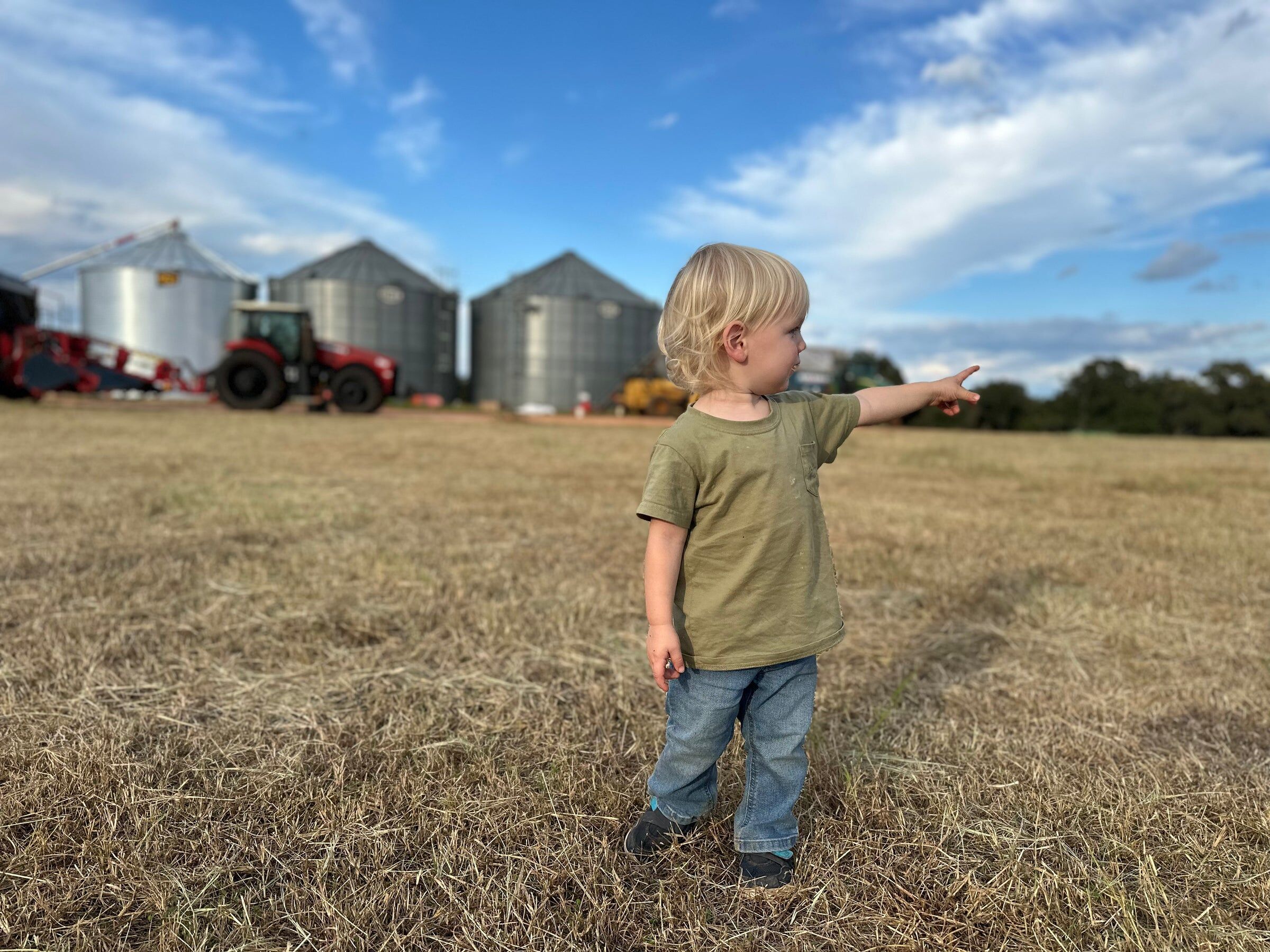 A young child stands in a cut field, pointing toward the farm’s grain bins and tractors.